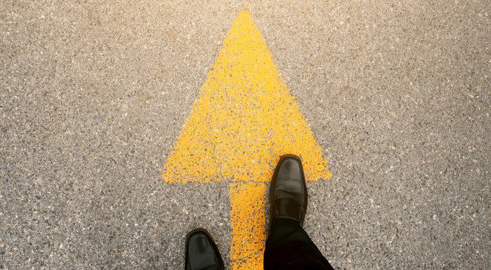 A photograph showing the point of view of a person looking down at their shoes whilst standing on a yellow arrow.