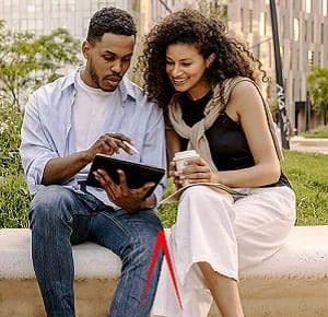A man holding a tablet computer, and a woman holding a disposable coffee cup, sitting outdoors on a stone bench in a park-like setting. They are looking at something on the tablet screen.
