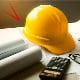 A yellow construction hard hat placed on a desk alongside rolled-up architectural plans and a black calculator, suggesting a work setting related to engineering, construction, or design.