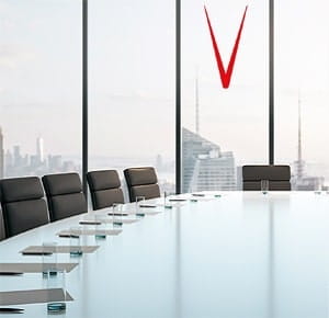 A modern conference room with a long glass-top table surrounded by black leather chairs. Large floor-to-ceiling windows in the background reveal a city skyline with tall buildings under a bright sky.