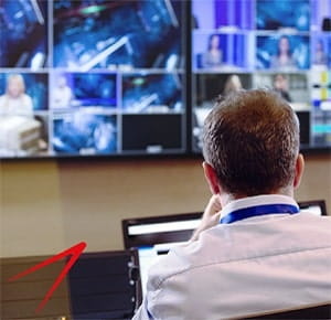A person seated at a workstation in front of multiple large screens displaying various video feeds and graphics, resembling a control room or monitoring setup. The individual is using a laptop, and the environment suggests a high-tech or broadcast operations setting.