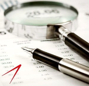 A close-up of a magnifying glass and a pen resting on a sheet of paper filled with printed numbers, likely financial or accounting data, symbolizing analysis, auditing, or detailed examination.