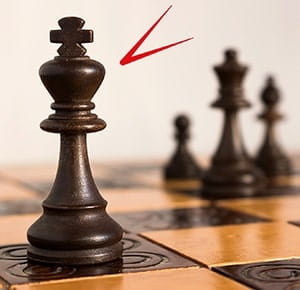 A close-up of a chessboard with a dark king piece prominently in the foreground, positioned on a light square. In the background, other dark chess pieces, including pawns, are slightly out of focus. The board features alternating light and dark squares, symbolizing strategy and decision-making.