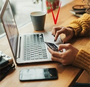 Hands typing on a laptop keyboard while holding a credit card, placed on a wooden table. A smartphone lies nearby, and a coffee cup is positioned in the background, suggesting an online payment or e-commerce activity in a casual workspace setting.