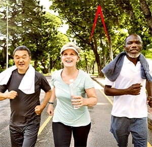 Three active, middle-aged people jogging together on a paved path lined with trees in an outdoor park setting. The background shows greenery and sunlight filtering through the trees.