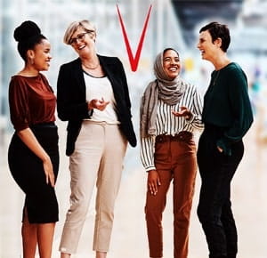Four women standing together indoors, engaged in conversation and smiling, with a blurred modern office background.