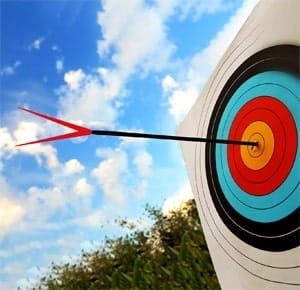 An archery target with concentric coloured rings, mounted outdoors against a backdrop of blue sky with scattered clouds. A single arrow is embedded in the target, striking near the center.