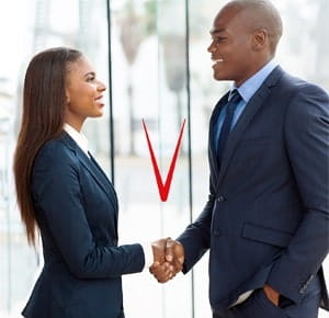 A man and a woman in formal business attire standing indoors near large glass windows, shaking hands in a professional setting.
