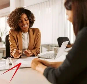 Two people seated at a desk in a bright office setting, engaged in a discussion. One person is holding a pen and gesturing toward documents on the table, while the other has a laptop open in front of them. The scene suggests a professional meeting or interview.
