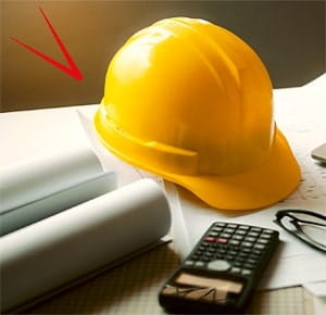A yellow construction hard hat placed on a desk alongside rolled-up architectural plans and a black calculator, suggesting a work setting related to engineering, construction, or design.