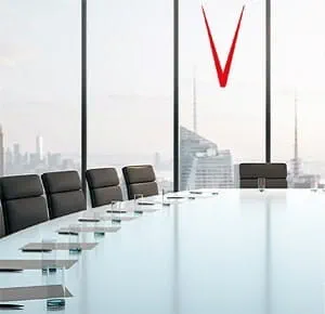 A modern conference room with a long glass-top table surrounded by black leather chairs. Large floor-to-ceiling windows in the background reveal a city skyline with tall buildings under a bright sky.