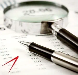A close-up of a magnifying glass and a pen resting on a sheet of paper filled with printed numbers, likely financial or accounting data, symbolizing analysis, auditing, or detailed examination.