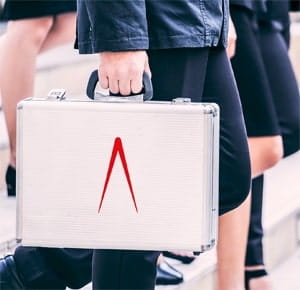 A person walking up steps while carrying a silver metal briefcase in one hand. The individual is dressed in dark clothing, and other people in similar attire are visible in the background, suggesting a professional or business setting.