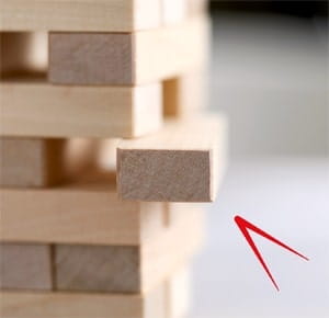 A wooden block tower game with one rectangular block partially pulled out from the stack, highlighting a moment of tension or strategy in gameplay. The blocks are light-coloured and arranged in alternating layers on a smooth surface.