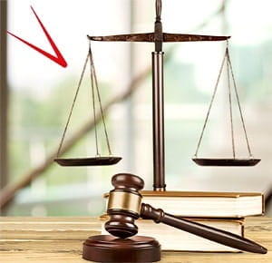 A wooden gavel resting on a table next to two stacked books, with a bronze balance scale in the background. The scene suggests themes of law, justice, or legal proceedings, set against a softly blurred indoor backdrop.