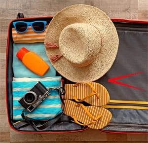 An open suitcase neatly packed with vacation essentials, including a straw hat, striped flip-flops, a folded beach towel, a pair of sunglasses, a bottle of sunscreen, and a vintage-style camera. The suitcase is placed on a wooden surface, suggesting preparation for a summer holiday or beach trip.