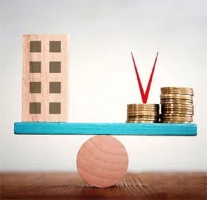 A wooden block shaped like a building and a stack of coins balanced on a blue plank resting on a round wooden base, symbolizing financial balance or property investment.