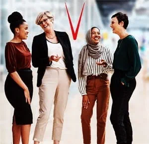 Four women standing together indoors, engaged in conversation and smiling, with a blurred modern office background.