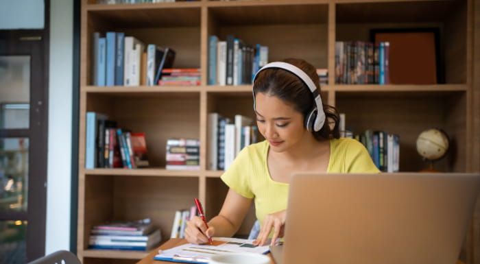 Young woman writing on paper and using a laptop wearing headphones