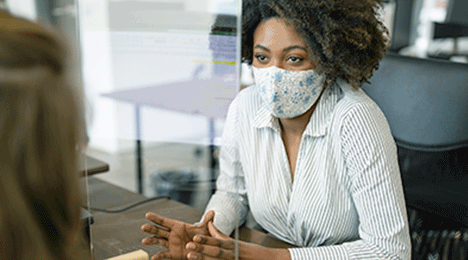 A woman at an office table wearing a face mask, talking to someone across the table from her.