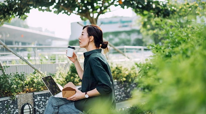 A person sitting outside on a bench with a laptop on their lap and a cup of coffee in one hand