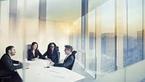 Four people sitting around a desk in an office with a cityscape visible through the windows