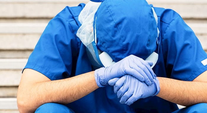 A medical professional in scrubs sitting outside on steps with their forehead resting on the hands