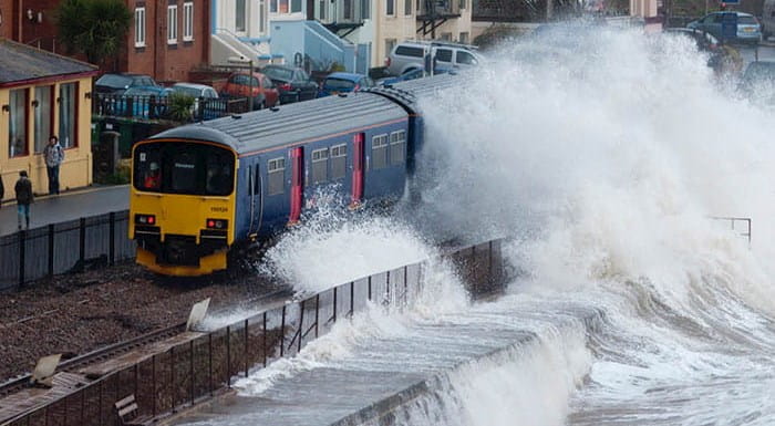 A big sea wave crashing over a train running along the coast.
