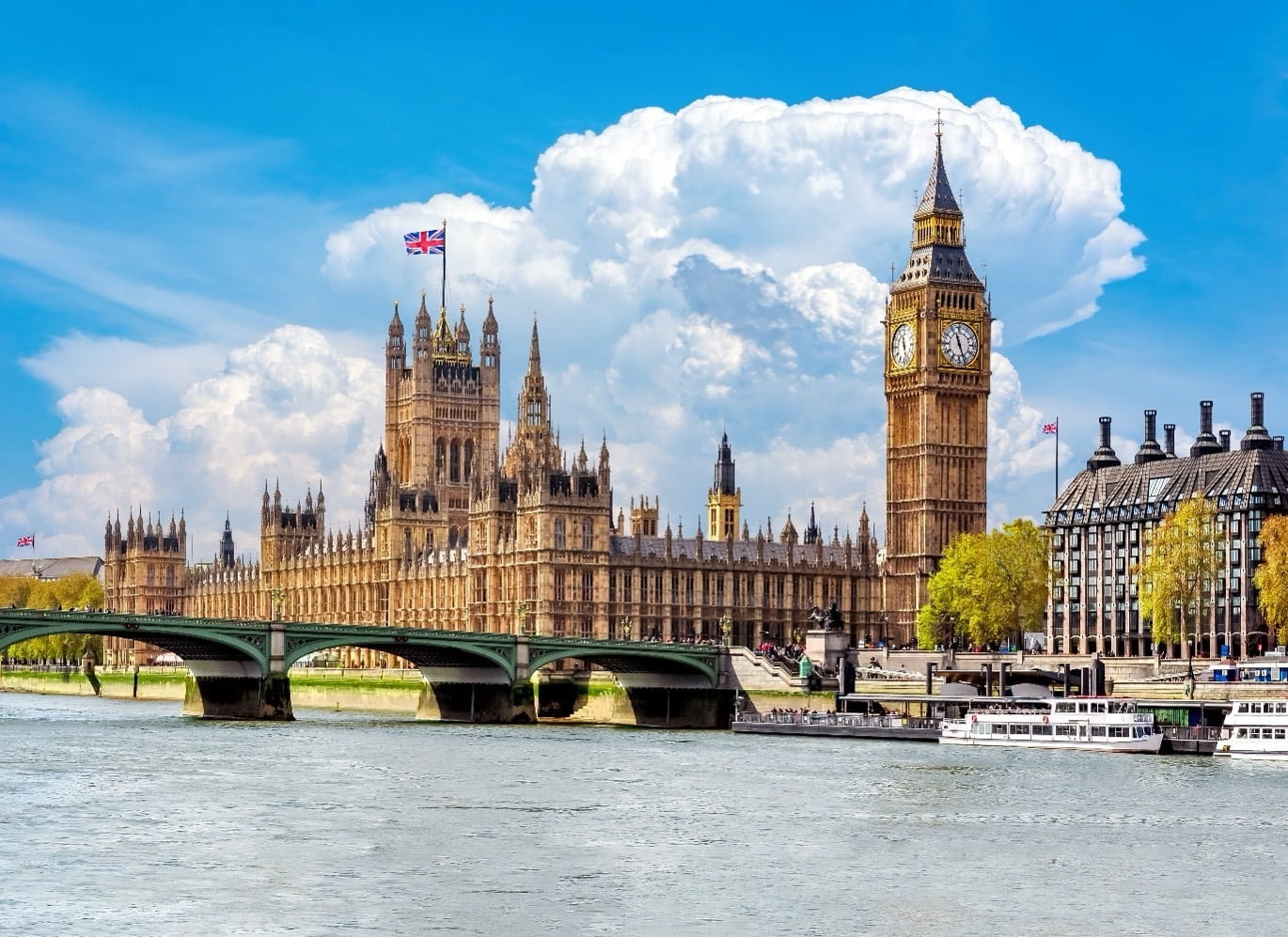 A wide view of the Houses of Parliament and Big Ben beside the River Thames under a bright blue sky with large clouds.