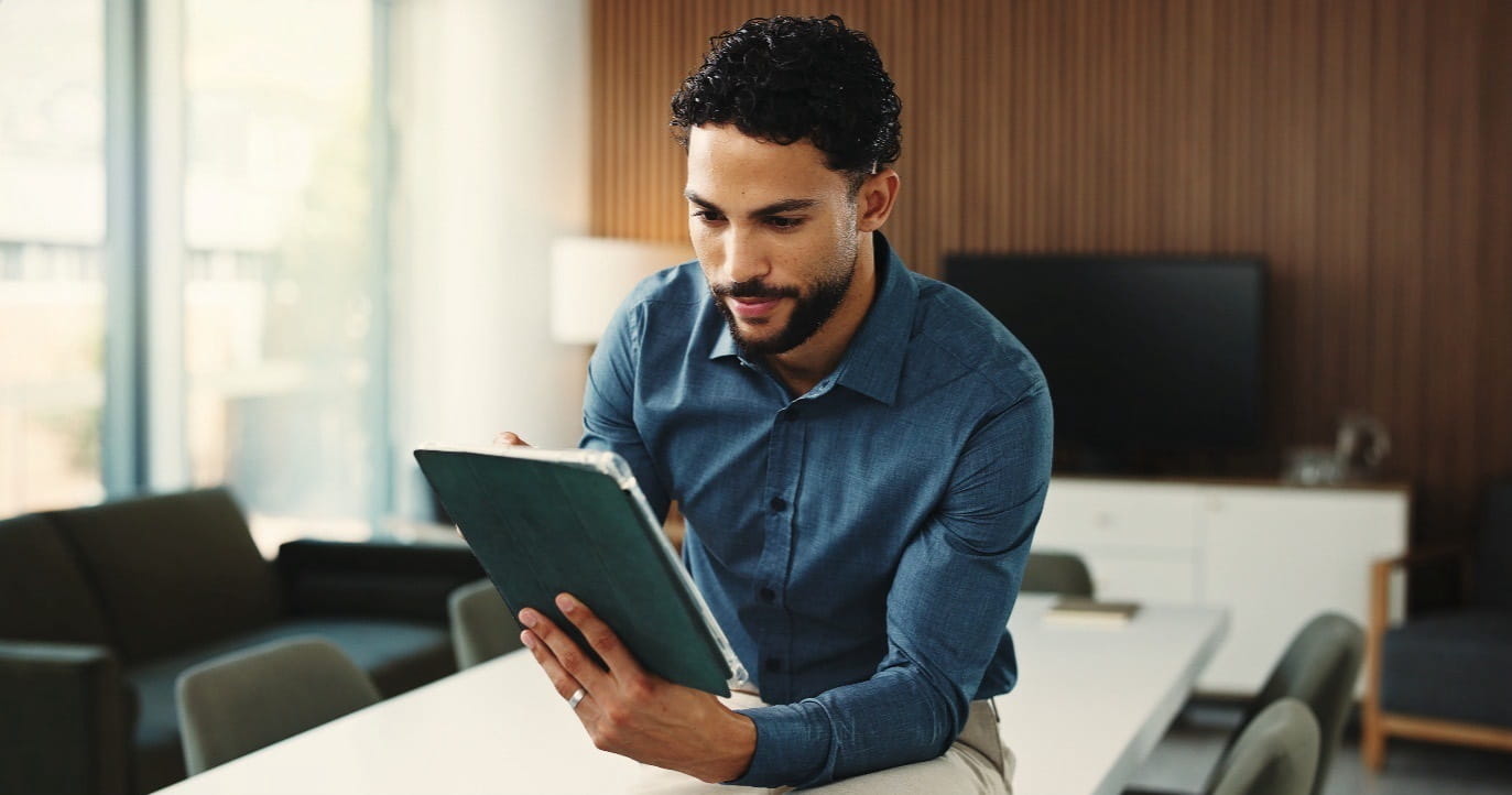 A person in a blue shirt sits on a table holding and reading from a tablet in a modern office space.