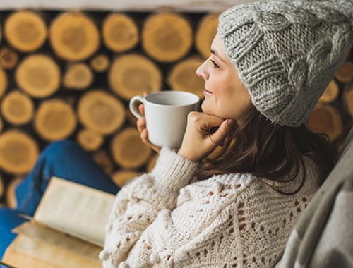 young woman knitted grey beanie hat cream jumper sweater mug hot drink book wood logs winter holiday