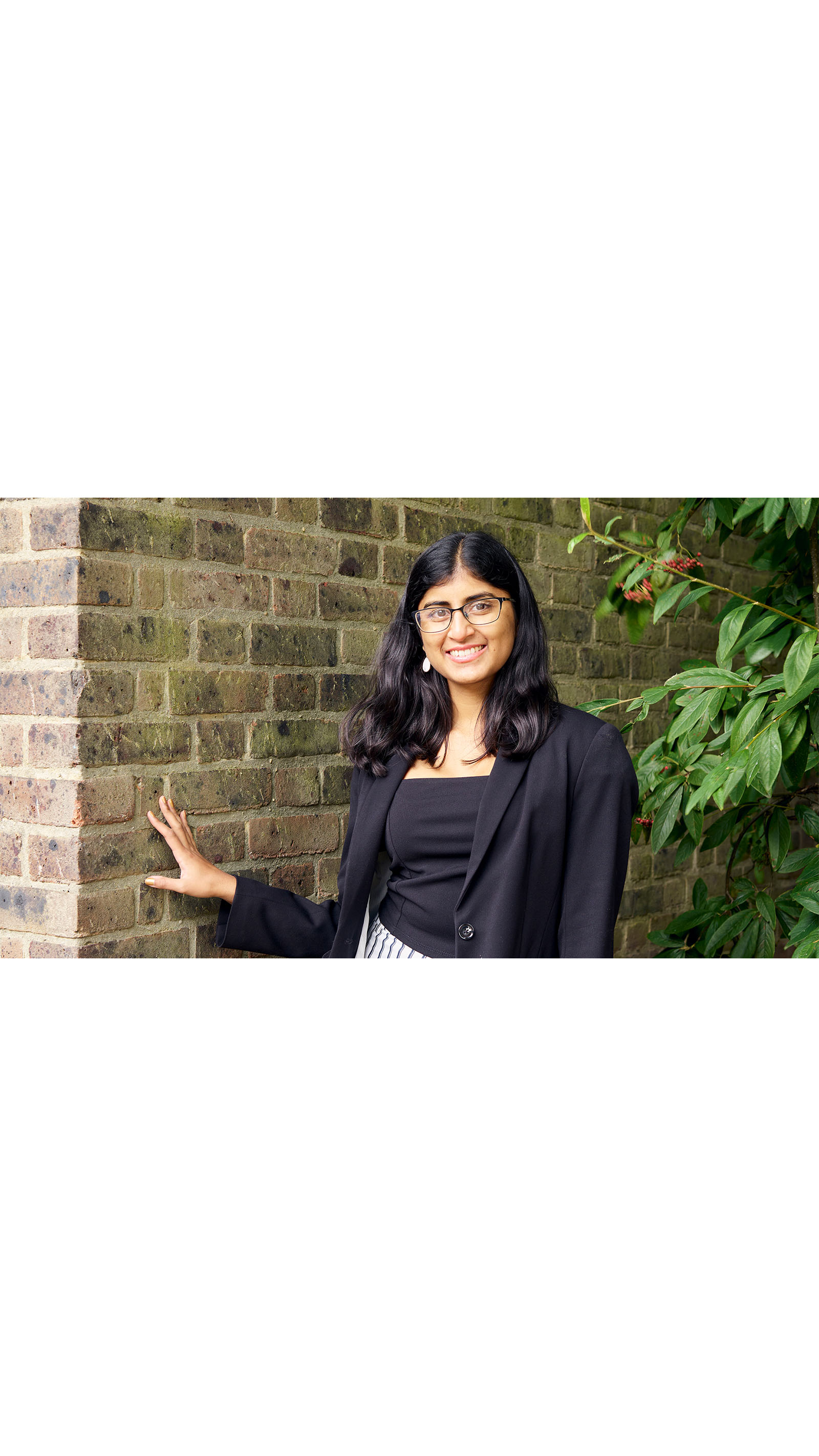 ACA student Ashwini Poopalasingham young asian woman standing in front of a brick wall and tree black clothing long hair