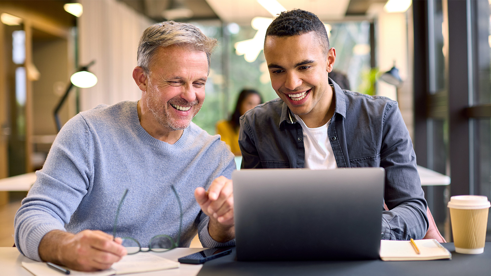 two men manager employee colleagues smiling looking at a laptop on a desk in an open office
