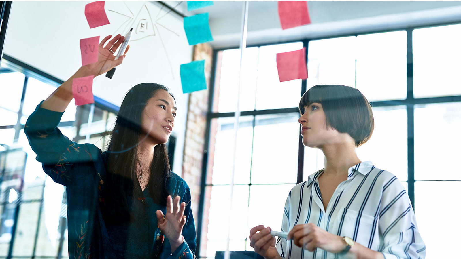 two women colleagues brainstorming meeting office large windows glass post it notes