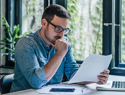 young man wearing glasses chin in hand looking at paperwork in an office desk laptop green plants