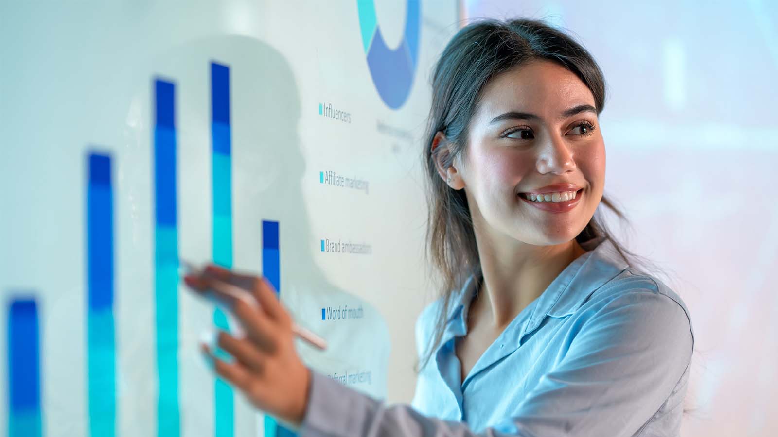young woman employee smiling working meeting room presenting pointing to a chart table on a digital screen