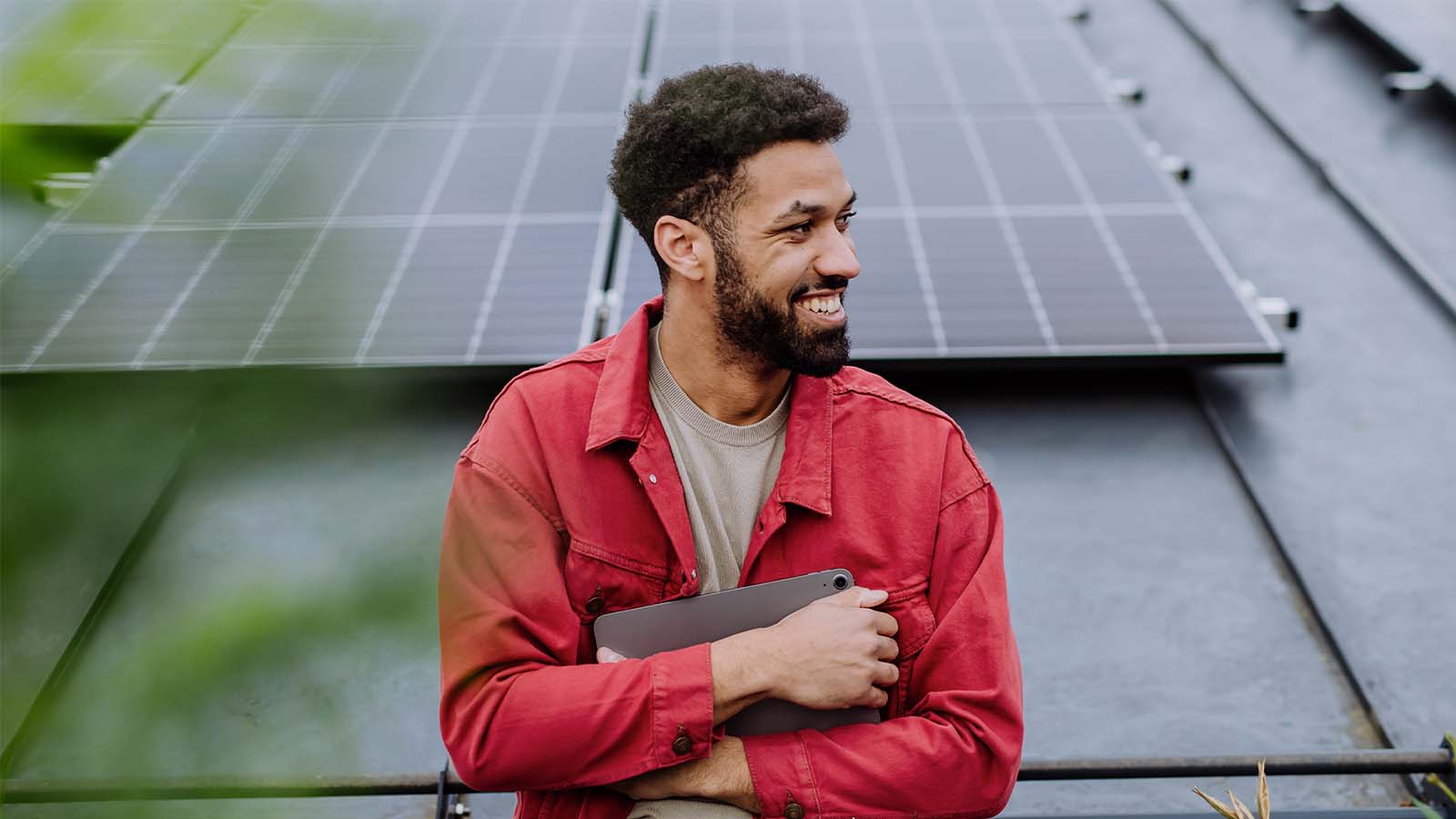 young man with a beard wearing a red shirt standing in from of solar panels on a rooftop sustainability