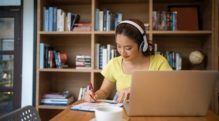 Person wearing headphones writing on papers at a table with an open laptop on it