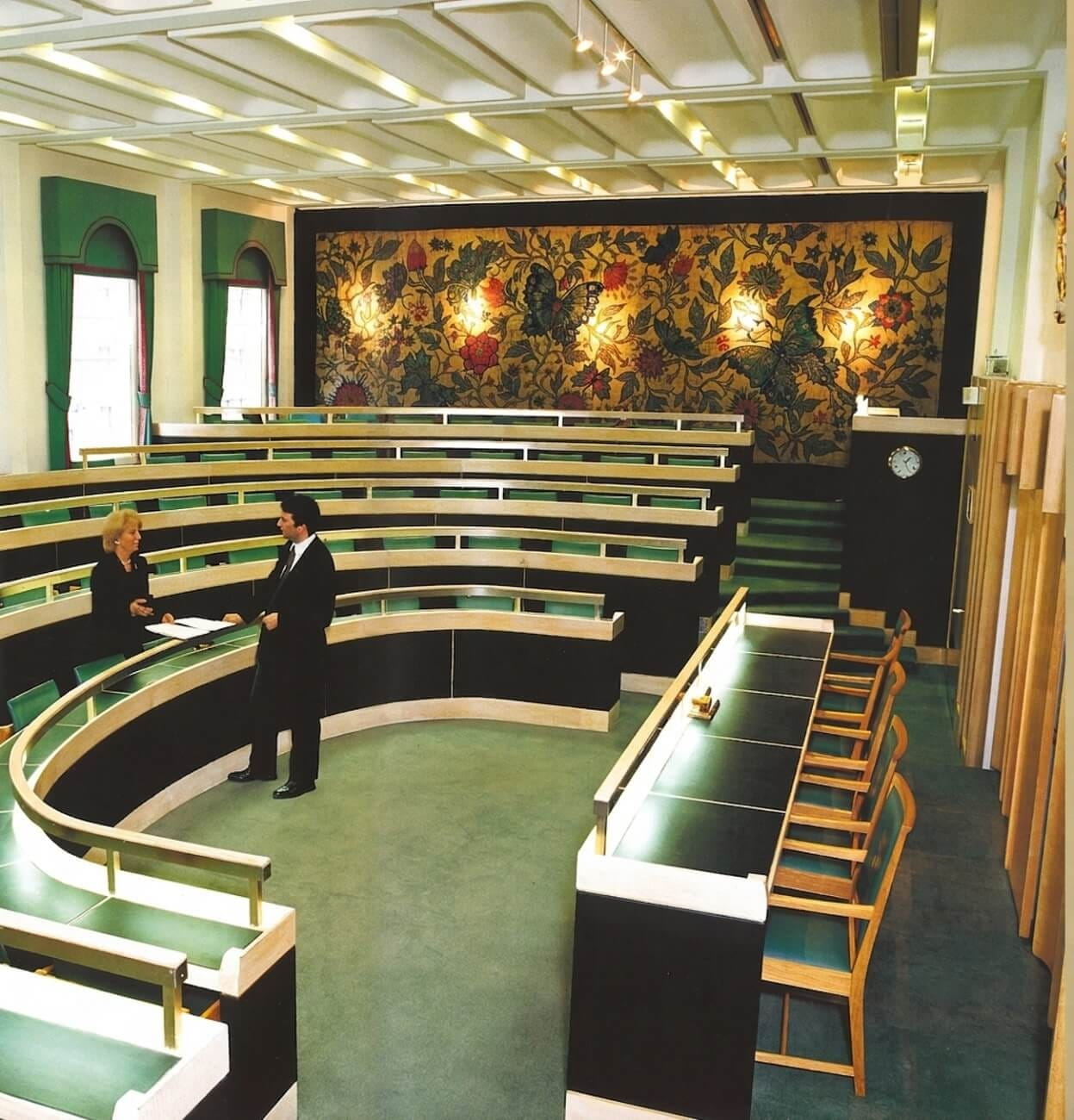 a small auditorium, with two standing people and a tapestry on the end wall