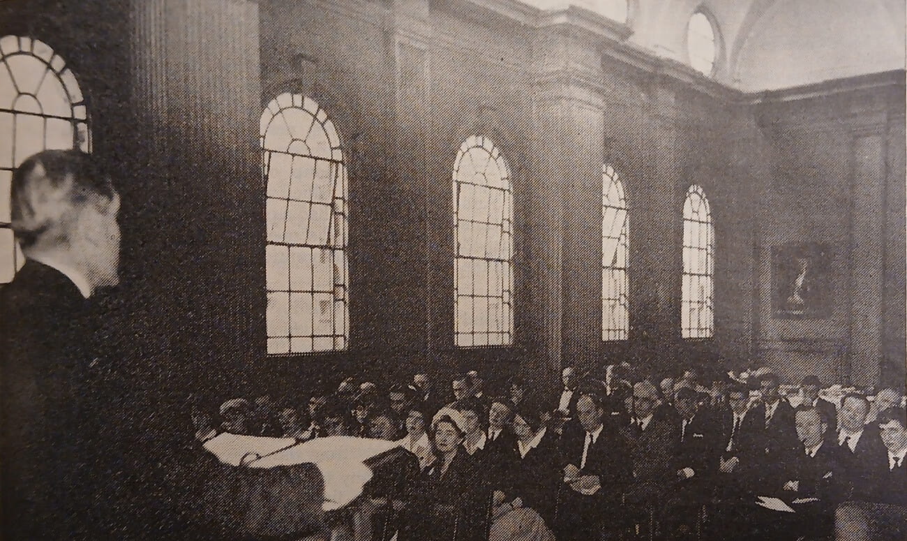A vintage picture of a man speaking to a group of people in a hall