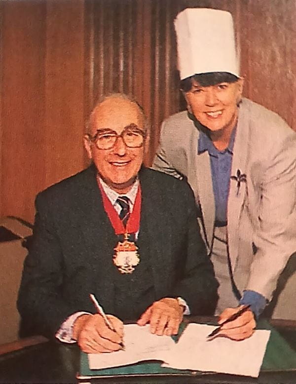 Man and woman signing a document, the woman has a chefs hat on, the man has a presidential chain around his neck