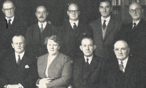 A black and white posed photoragph of a group of man and one woman, all formally dressed