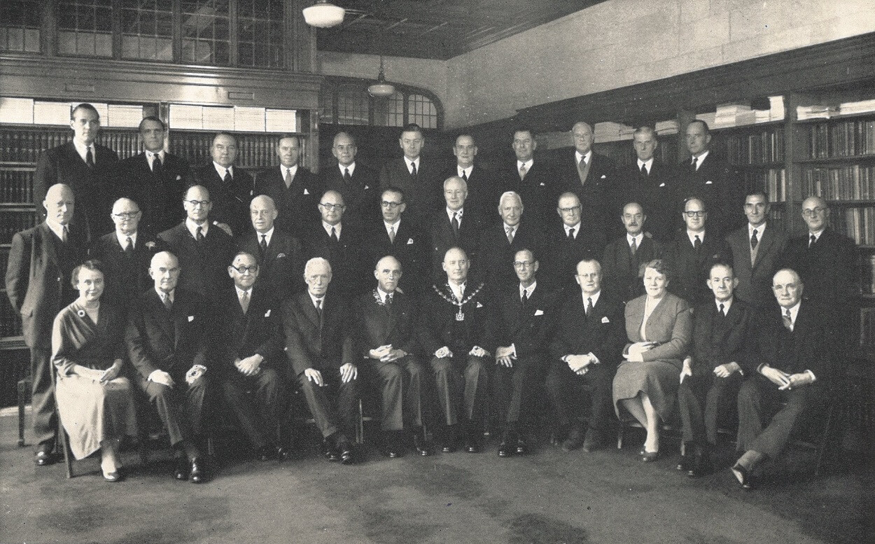 A black and white posed team photograph of a group of man and two woman, all formally dressed