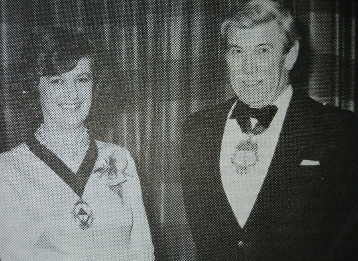 A vintage photograph of a smiling young woman with a badge of office on a ribbon around her neck with an older man in formal dress, also with a badge of office around his neck