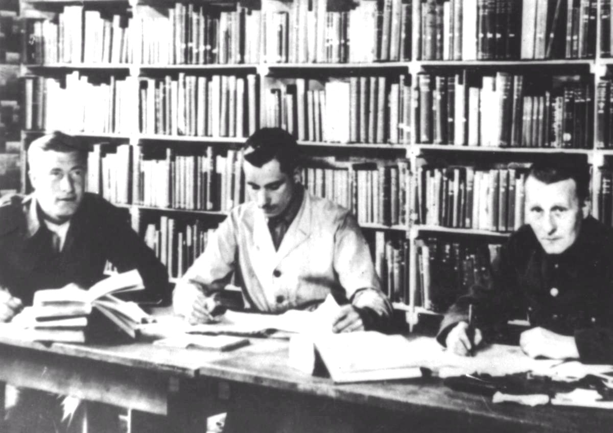 Vintage photograph of a library in a prisoner of war camp showing books on shelves behind 3 men looking at books on a table