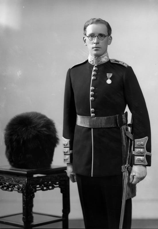 3 quarter length shot of a man in military dress uniform with a medal pinned to chest. A bearskin hat is on a table to his left
