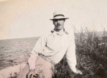 Black and white photograph if William Harris-Burland sitting on a beach. He is dressed in a white shirt and light trousers and is wearing a hat.