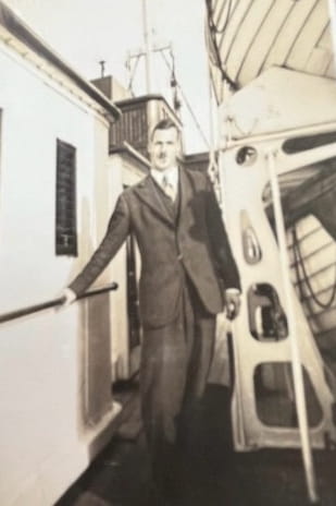 Black and white photograph of William Harris-Burland in a three-piece suit, stood on the deck of a ship next to the wooden lifeboats.