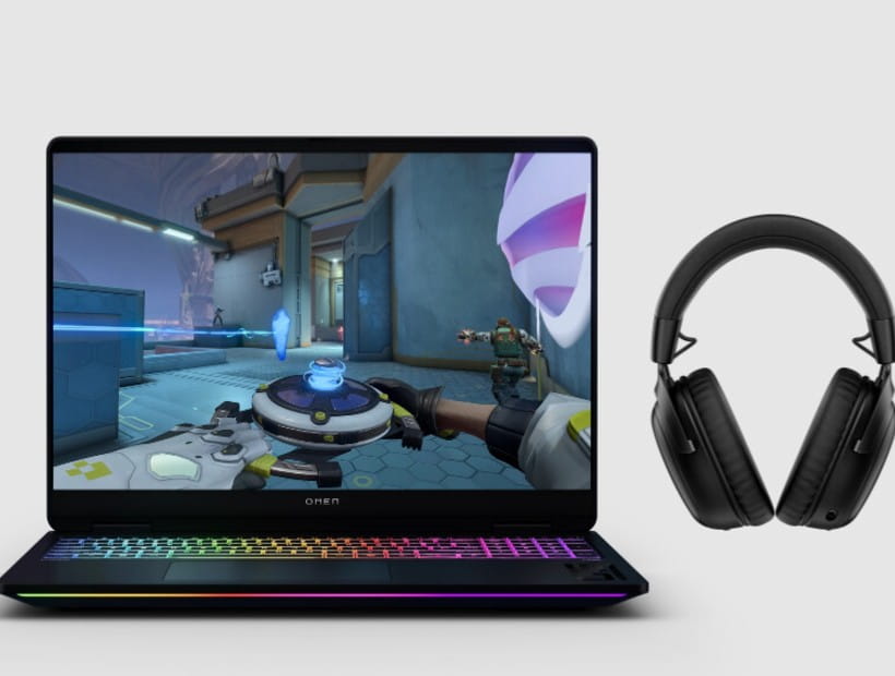 A gaming laptop displaying a first-person shooter game sits beside a pair of black over‑ear headphones on a plain background.