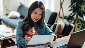 A person working at a table with a laptop and papers, and a baby on their lap